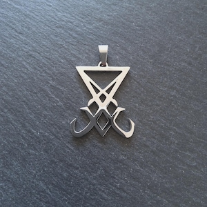 Può includere: Un ciondolo color argento raffigurante il Sigillo di Lucifero. Il ciondolo ha una parte superiore triangolare con un design stilizzato sottostante e due forme curve nella parte inferiore. Il ciondolo è mostrato su una superficie grigio scuro.