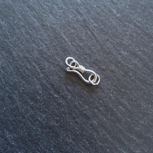 Può includere: Una piccola chiusura per gioielli color argento con un design a gancio e occhiello. La chiusura è in metallo e ha un gancio ad anello su un'estremità e un occhiello ad anello sull'altra, collegati da una sezione di filo avvolto. La chiusura è su una superficie grigio scuro strutturata.