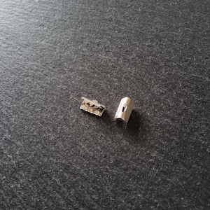 Puede incluir: Dos pequeños cierres metálicos plateados. Uno es rectangular con una serie de pequeños recortes decorativos a lo largo de un borde. El otro tiene forma cilíndrica redondeada con una pequeña abertura.
