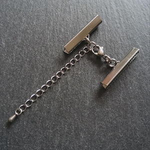 May include: Silver-coloured metal clasp featuring two rectangular bars and a chain extension. The clasp has a lobster claw closure, suitable for jewellery making or crafts. Set against a dark grey, textured surface.