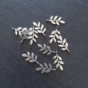 Puede incluir: Varios dijes plateados con forma de hojas dispersos sobre una superficie oscura. Cada dije presenta un diseño detallado de hoja, ideal para la creación de joyas o proyectos de artesanía. Los dijes son pequeños y delicados.