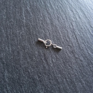May include: A small, silver-coloured jewellery clasp. The clasp comprises a ring, a small bar, and a connecting bar with a small loop. The clasp is designed to secure jewellery pieces such as necklaces or bracelets.