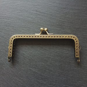 May include: An antique brass purse frame, rectangular in shape, with a decorative perforated pattern along the top. The frame features a clasp with two small, round knobs and loops for attaching to a purse.