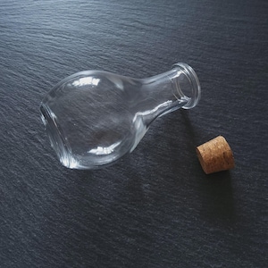 May include: A clear glass bottle with a rounded body and a narrow neck, lying on its side. A cork stopper is positioned nearby. The background is a dark, textured surface.