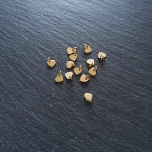 Può includere: Una collezione di piccoli ciondoli a forma di cuore color oro sparsi su una superficie scura e strutturata. Ogni ciondolo ha un piccolo anello nella parte superiore, suggerendo che sono progettati per la creazione di gioielli o l'artigianato.