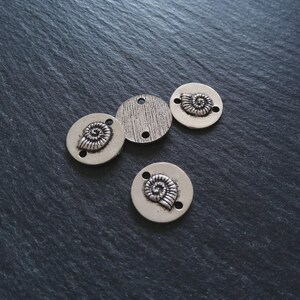 May include: Four silver-coloured metal charms. Three charms are round with a raised spiral design and two holes. One charm is round with a textured surface and two holes. The charms are on a dark grey surface.