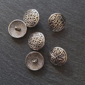 Puede incluir: Botones de metal plateado con un diseño de nudo celta. Los botones son redondos con un patrón detallado en la parte delantera y un vástago en la parte posterior. Los botones están dispersos sobre una superficie gris oscuro.