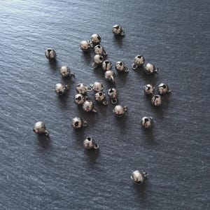 May include: A scattering of small, shiny silver bells on a dark grey surface. Each bell has a small loop at the top for attaching. The bells are round with a slit opening.