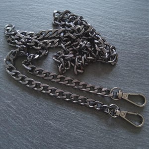 Puede incluir: Una cadena de metal gris plomo con dos cierres de mosquet&oacute;n. La cadena est&aacute; hecha de eslabones ovalados entrelazados. La cadena est&aacute; sobre una superficie gris oscuro.