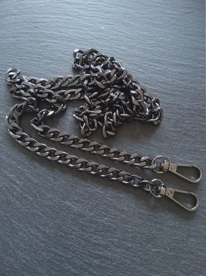 Puede incluir: Una cadena de metal gris plomo con dos cierres de mosquet&oacute;n. La cadena est&aacute; hecha de eslabones ovalados entrelazados. La cadena est&aacute; sobre una superficie gris oscuro.