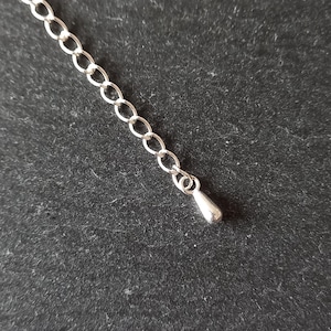 May include: A silver-colored chain extender with a teardrop-shaped charm at the end. The chain is composed of small, interconnected oval links. The background is a dark, textured surface.