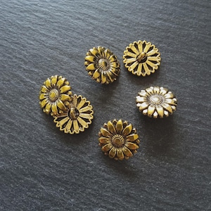 May include: Six antique gold-coloured daisy-shaped buttons are displayed on a dark grey, textured surface. The buttons have detailed petal and centre designs, suggesting a vintage or handcrafted aesthetic. The buttons are likely for sewing or craft projects.