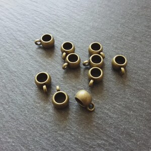 May include: A selection of small, bronze-coloured cup charms with tiny handles. The charms are scattered on a dark grey surface, with the cups facing in various directions. These miniature cups are ideal for jewellery making or crafting projects.