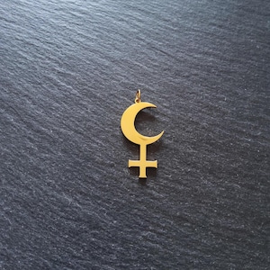 Può includere: Un ciondolo dorato raffigurante una mezzaluna sopra un simbolo a forma di croce. Il ciondolo è sospeso a un piccolo anello, suggerendo che sia progettato per essere indossato come gioiello. Lo sfondo è una superficie scura e strutturata.