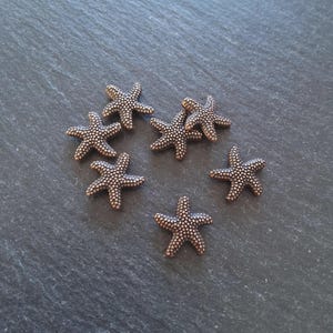 May include: Seven bronze starfish on a dark grey surface. Each starfish has a textured surface with small, evenly spaced dots. The starfish are of similar size and shape, creating a decorative arrangement.