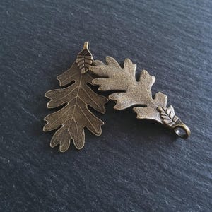 May include: Two bronze-colored oak leaf pendants. Each leaf has detailed veining and a small loop at the top for hanging. The leaves are slightly different in position, with one overlapping the other. The background is a dark, textured surface.