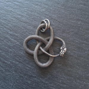 May include: A silver snake pendant with a textured, scale-like pattern. The snake is coiled in a knot shape, with a detailed head and two rings at the top. The pendant is displayed on a dark grey surface.