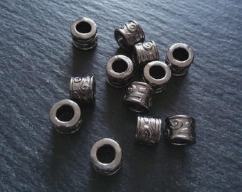 May include: A selection of dark grey metal beads, cylindrical with a central hole. Some beads have an etched swirl pattern. The beads are scattered on a dark grey surface, ideal for jewellery making.