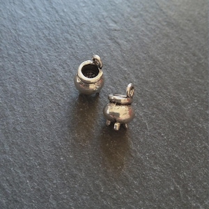 May include: Two small, silver-coloured cauldron charms. Each charm has a rounded body, three small feet, and a handle. One cauldron has its lid open. The charms are displayed on a dark grey, textured surface.