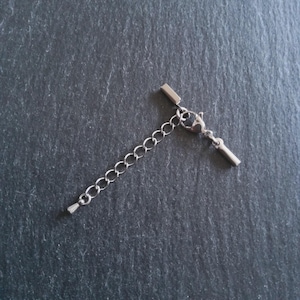 May include: A silver-colored necklace extender with a chain and a T-bar clasp. The chain has small, oval links. The extender is designed to add length to necklaces or bracelets. The background is a dark gray, textured surface.