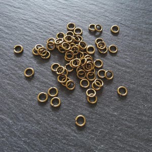 Puede incluir: Un conjunto de pequeños anillos de latón dispersos sobre una superficie oscura y texturizada. Los anillos son circulares, con una pequeña abertura, y varían en cantidad. Ideal para la creación de joyas o proyectos de artesanía.