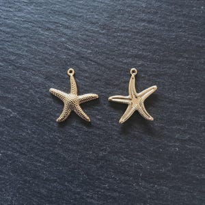 May include: Two gold starfish charms with textured surfaces and small loops for hanging. The charms are displayed on a dark grey background, ideal for jewellery making or craft projects.