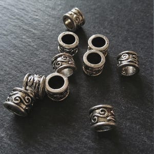 Op de afbeelding: Een verzameling zilverkleurige metalen kralen met een antieke afwerking. Elke kraal heeft een cilindrische vorm met ingewikkelde wervelpatronen. De kralen zijn verspreid over een donker, getextureerd oppervlak, wat wijst op een ambachts- of sieraden thema.