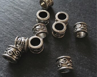 May include: Silver-coloured metal beads with an antique finish. Each cylindrical bead has detailed swirling patterns. The beads are scattered on a dark, textured surface, suggesting a craft or jewellery-making theme.