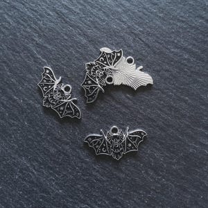 Może przedstawiać: Trzy srebrne zawieszki w kształcie nietoperzy ze skomplikowanymi detalami, w tym akcentami gwiazd i księżyca. Zawieszki mają pętlę do mocowania do biżuterii lub innych rękodzieł. Nietoperze są prezentowane na ciemnej, teksturowanej powierzchni.