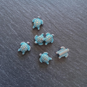 May include: A collection of small turtle figurines in blue and brown hues, displayed on a dark grey surface. The turtles feature intricate shell designs and appear to be made of metal. These could be used for jewellery or craft projects.