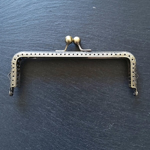 Puede incluir: Un marco de bolso de estilo vintage con acabado bronce. El marco tiene forma rectangular con detalles decorativos en los bordes y un cierre de broche con dos pequeños pomos redondos. Tiene pequeños agujeros para coser.