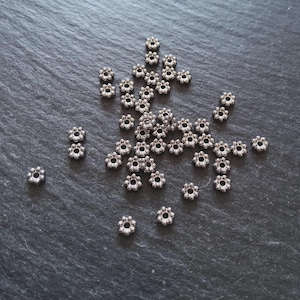 Op de afbeelding: Kleine, zilverkleurige metalen bloemkralen verspreid op een donkergrijze ondergrond. Elke kraal heeft een bloemmotief met meerdere bloemblaadjes en een centraal gat. Geschikt voor het maken van sieraden of knutselen.