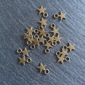 Puede incluir: Un conjunto de pequeños dijes de estrella de color bronce con anillas adjuntas. Los dijes están dispersos sobre una superficie texturizada de color gris oscuro. Cada estrella tiene cinco puntas y un pequeño lazo.