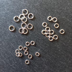 May include: A collection of small, circular silver-toned metal jump rings arranged in three distinct clusters. Each individual ring features a single open seam, allowing for connection and assembly in jewelry making or other craft projects.