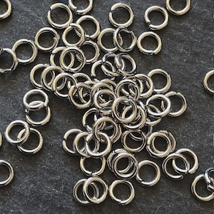 Puede incluir: Primer plano de numerosos anillos de salto plateados, esparcidos sobre una superficie gris oscuro. Los anillos son circulares con una pequeña abertura, ideales para la fabricación de joyas o manualidades. La luz resalta su brillo metálico.