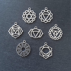 Puede incluir: Siete colgantes de chakra plateados con varios diseños geométricos, incluyendo estrellas, triángulos y patrones florales. Cada colgante tiene un pequeño lazo en la parte superior para sujetarlo a una cadena o cordón. Los colgantes se muestran sobre una superficie gris oscuro.