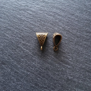 May include: Two small, antique brass-colored jewelry findings. One is a triangle shape with an ornate design, and the other is a rounded shape with a dark interior. Both have small loops for attaching to jewelry.