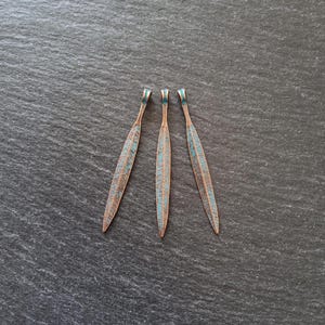 May include: Three copper-coloured feather pendants with turquoise accents. Each pendant has a long, slender shape with detailed veining and a small loop at the top. The pendants are arranged on a dark grey surface.