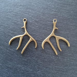 May include: Two bronze-coloured antler-shaped charms with a loop at the top for jewellery making. The charms have a detailed, organic design with multiple curved tines. They are displayed on a dark grey textured surface.