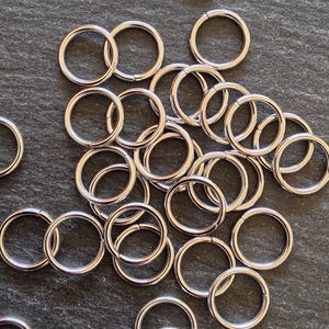 Pode incluir: Uma coleção de anéis de metal prateados brilhantes espalhados sobre uma superfície cinza escuro. Os anéis são circulares com uma pequena abertura, sugerindo que são articulados ou projetados para abrir e fechar. A iluminação destaca as superfícies lisas e reflexivas dos anéis.