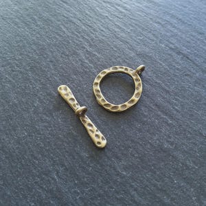 Puede incluir: Un conjunto de cierre de palanca de color bronce, que presenta una barra texturizada y un anillo martillado con un pequeño lazo. El cierre está diseñado para la fabricación de joyas y se muestra sobre una superficie gris oscuro.