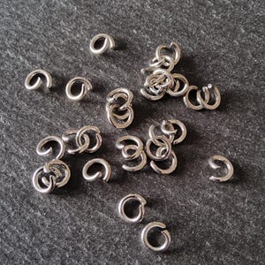 May include: A scattering of small, shiny, silver jump rings on a dark grey textured surface. The jump rings are open, ready for use in jewellery making or other crafts. The image is well-lit, showing the metallic sheen of the rings.