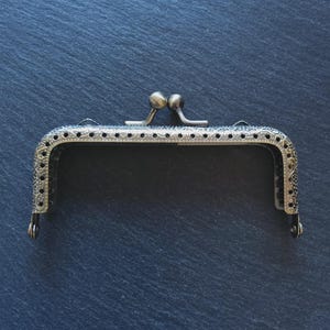 Puede incluir: Un marco de bolso antiguo en tono bronce con un patrón floral decorativo. El marco tiene un cierre de beso con dos pequeños cierres en forma de bola. El marco está diseñado para ser cosido en un bolso o bolsa.