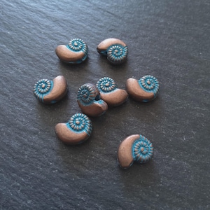 May include: A collection of copper-toned, shell-shaped beads with turquoise blue spiral patterns. The beads are displayed on a dark grey surface. These decorative beads are suitable for crafting jewellery and other creative projects. Each bead is approximately 1.5 cm in length.