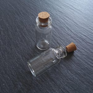 May include: Two small, clear glass bottles with cork stoppers. One bottle is upright, and the other is lying on its side. The bottles are on a dark grey, textured surface.