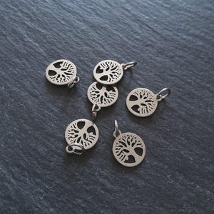 Op de afbeelding: Zes zilverkleurige levensboom bedels met een cirkelvormig ontwerp. Elke bedel heeft een kleine lus om aan sieraden te bevestigen. De bedels worden gepresenteerd op een donkergrijze, getextureerde ondergrond.