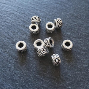 Puede incluir: Un conjunto de cuentas de metal plateado con intrincados diseños geométricos. Las cuentas cilíndricas tienen un agujero en el centro para ensartar. Se muestran sobre una superficie gris oscuro.
