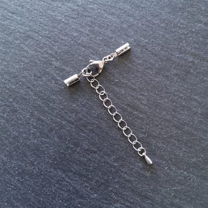 May include: A silver-colored necklace extender with a lobster clasp and a series of small, connected rings. The extender includes a small, teardrop-shaped charm at the end. The metal components are set against a dark, textured background.