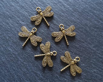 May include: Five bronze-colored dragonfly charms with detailed wing patterns and small loops for attaching to jewelry or crafts. The dragonflies are arranged on a dark, textured surface.