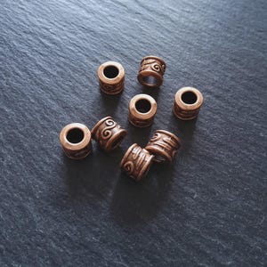 May include: A collection of eight bronze-colored metal beads. Each bead is cylindrical with a central hole and decorative etched swirl patterns. The beads are scattered on a dark gray surface.